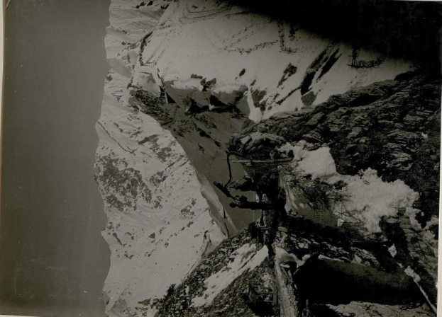 Schwarzes und weißes Foto eines Mannes in Kletterausrüstung, der einen schneebedeckten Berg mit dem Gipfel des Mount Everest im Hintergrund hinaufsteigt.
