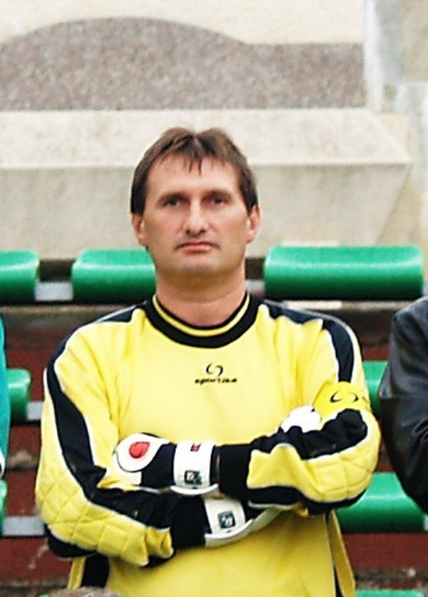 Ein Torwart in gelbem Trikot steht neben einem anderen Mann in einer schwarzen Jacke, mit grünen Stühlen und einer Wand im Hintergrund.