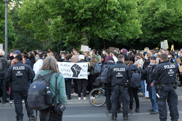 Eine große Gruppe von Menschen mit Schildern und Mützen und Taschen steht an einer Straßenkreuzung während einer Black Lives Matter Demonstration in Berlin, mit einem Fahrrad im Vordergrund und Bäumen und einem Pfahl im Hintergrund.
