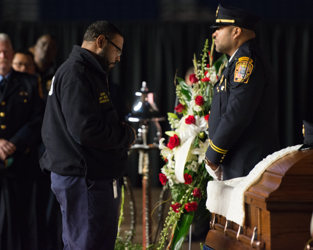 Zwei Polizisten in Uniform stehen feierlich neben einem mit Blumen geschmückten Sarg, mit einigen Personen im Hintergrund.