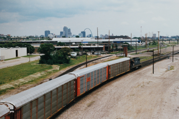Güterzug auf Schienen neben einer Stadtlandschaft mit Strommasten, Gebäuden, Bäumen, Gras und einem bewölkten Himmel.