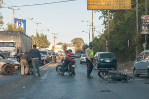 Eine Gruppe von Menschen steht um ein verunglücktes Motorrad auf der Straßenseite mit mehreren Fahrzeugen, darunter ein Lastwagen, und Hintergrundelementen wie Bäumen, Pfosten, Lampen, Schildern und dem Himmel.