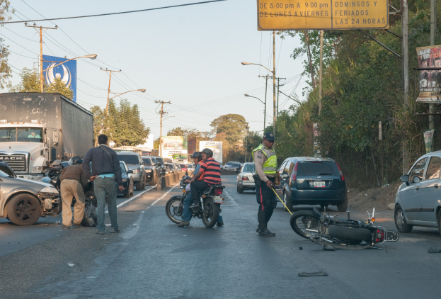 Eine Gruppe von Menschen steht um ein verunglücktes Motorrad auf der Straßenseite mit mehreren Fahrzeugen, darunter ein Lastwagen, und Hintergrundelementen wie Bäumen, Pfosten, Lampen, Schildern und dem Himmel.