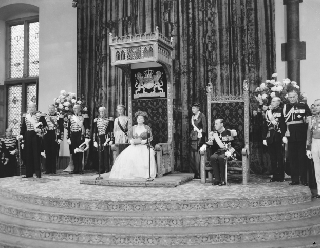 Königin Elizabeth II. und Prinz Philip, Duke of Edinburgh, auf ihrer Hochzeit sitzend, mit der Queen in einem weißen Kleid und einem Blumenstrauß, umgeben von stehenden Begleitern mit Blumen, in einem Raum mit einem Fenster und einem Vorhang.