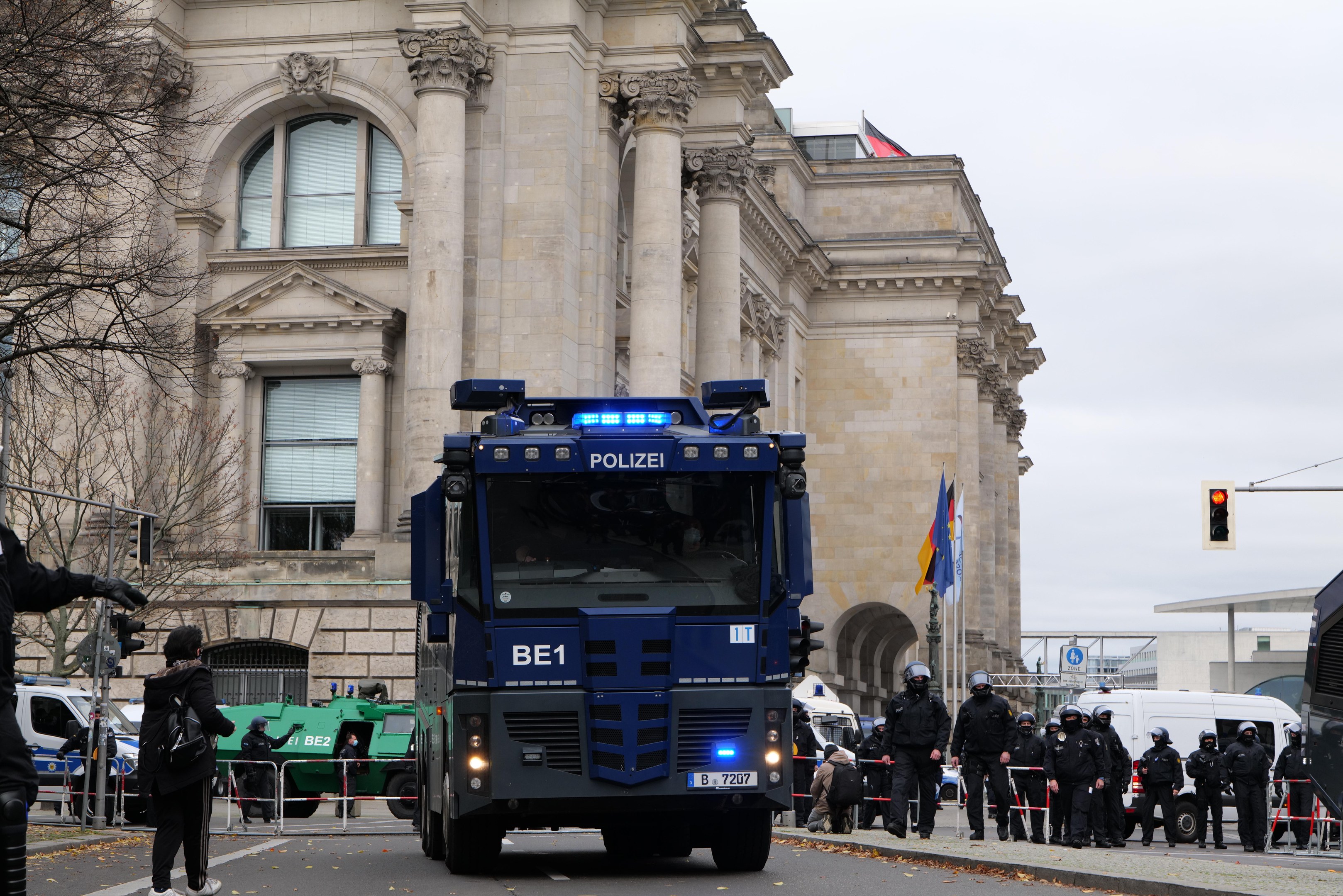 Eine Gruppe von Polizisten steht vor einem großen Gebäude mit architektonischen Details, Fahrzeugen auf der Straße und einer Person mit einer Kamera links. Im Hintergrund sind Bäume, Ampeln, Fahnenmasten und ein klarer blauer Himmel zu sehen.