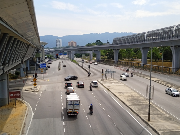 Eine stark befahrene Autobahn verläuft parallel zu einer Brücke, gesäumt von Straßenlaternen, Ampeln, Schildern, Bäumen, Gebäuden, Hügeln und einem bewölkten Himmel.