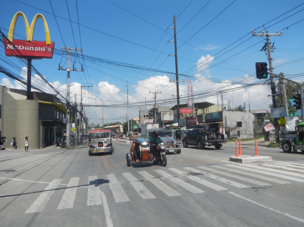 Vielbefahrene Straßenszene mit einem McDonald's-Schild, Fahrzeugen, Verkehrskegeln, Strommasten, Verkehrsampeln, Gebäuden, Schildern, Bäumen und einem bewölkten Himmel.