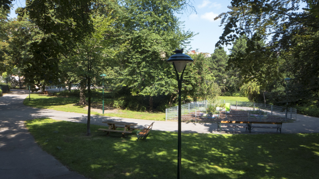 Ein Park mit einer Straße auf der linken Seite, Grasboden, Bänken, einer Laterne, einem Zaun, Pflanzen, Bäumen und einem bewölkten Himmel, mit einem Picknicktisch in der Mitte, der von Bäumen umgeben ist.