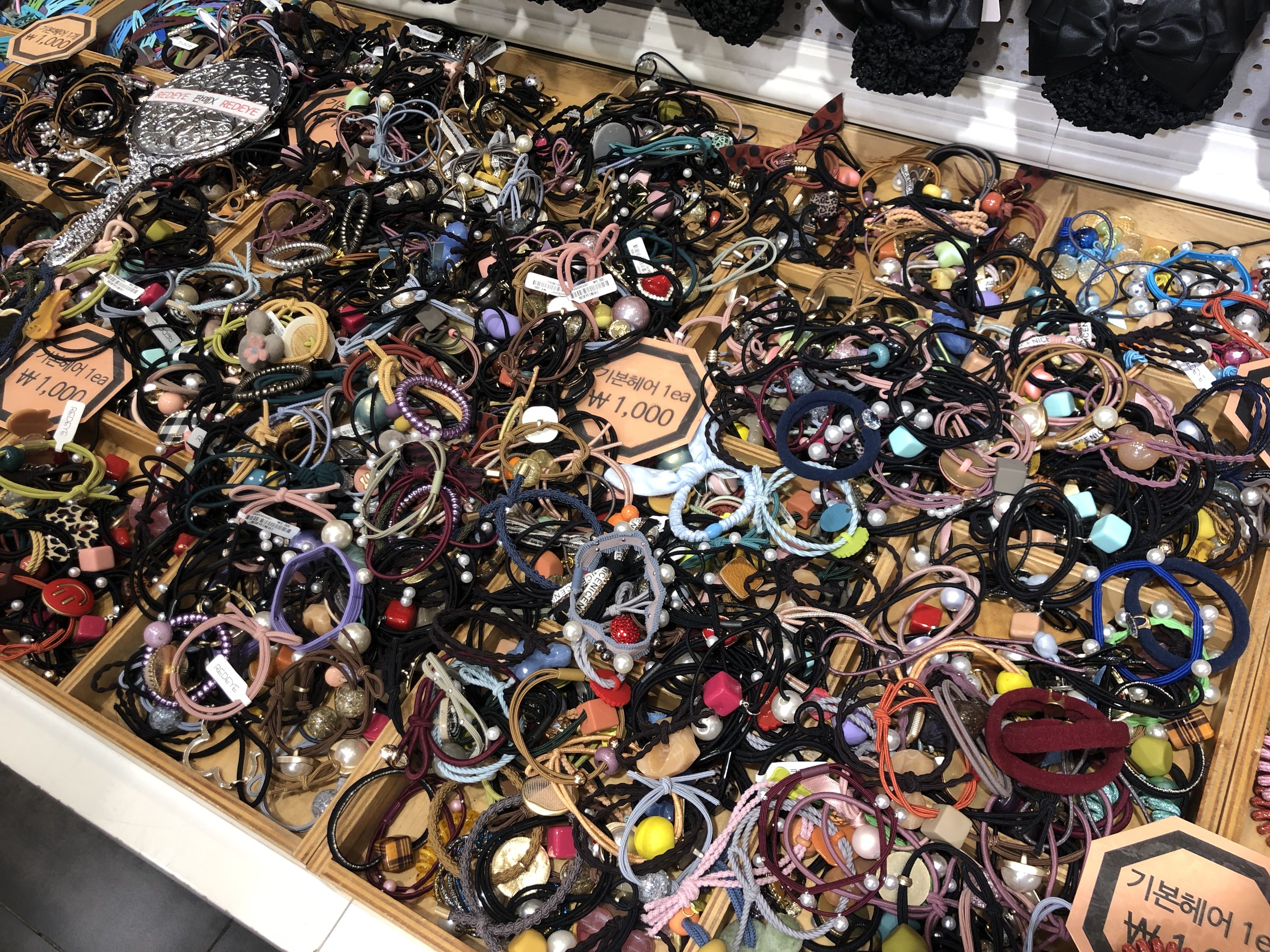 Vitrine voller verschiedener Schmuckstücke wie Armbänder, Ketten und Preisschilder, mit Kleidung darüber, die einen Marktplatz andeutet.