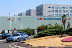 Krankenhausgebäude mit parkenden Autos, umgeben von städtischen Elementen einschließlich Gebäuden, Straßenmöbeln, Fahrzeugen, Grünflächen und einem klaren Himmel.