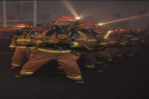 Feuerwehrleute in Gasmasken und Helmen stehen auf einer Straße und halten Feuerlöscher, mit einem Gebäude und Lichtern im Hintergrund und Text unten.