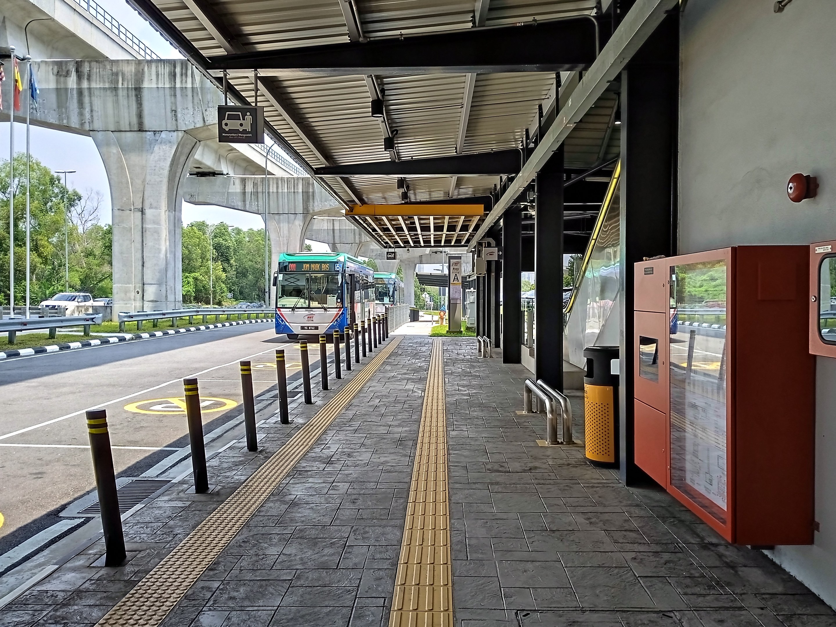 Eine Bushaltestelle mit einem Bus auf der Straße, Poller, Mülltonnen, ein Schuppen, Säulen, ein Schild, Fahnenmasten mit Fahnen, eine Brücke, Bäume und einen Himmel im Hintergrund.