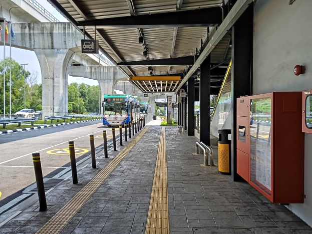 Eine Bushaltestelle mit einem Bus auf der Straße, Poller, Mülltonnen, ein Schuppen, Säulen, ein Schild, Fahnenmasten mit Fahnen, eine Brücke, Bäume und einen Himmel im Hintergrund.
