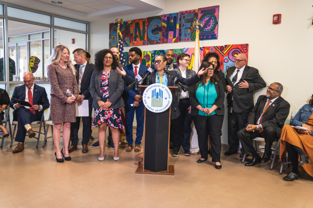 Eine Gruppe von Menschen, die vor einem Podium mit Mikrofon und einer Tafel mit der Aufschrift "Sancho-Stadtrat tritt dem Stadtrat bei" stehen und sitzen, mit Flaggen, einer Tafel, einem Feuerlöscher und Deckenleuchten an der Rückwand.