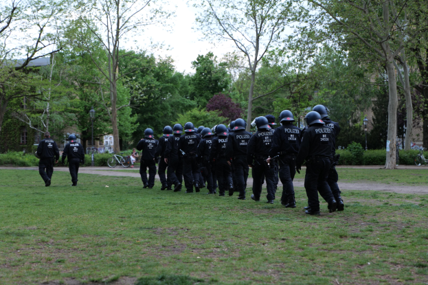Eine Gruppe von Polizisten in schwarzen Uniformen und Helmen, die über ein grünes Feld gehen, mit Fahrrädern, Laternenmasten, Pflanzen, Bäumen, Gebäuden und einem klaren blauen Himmel im Hintergrund.