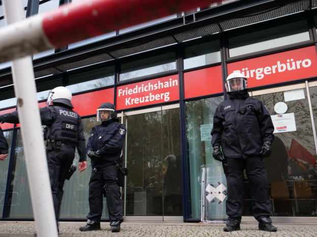 Polizisten in Uniform vor einem Glaswandgebäude mit angeklebten Papieren, einem Pfahl links und Hintergrundschildern mit der Aufschrift "Friedrichshain Kreuzberg."