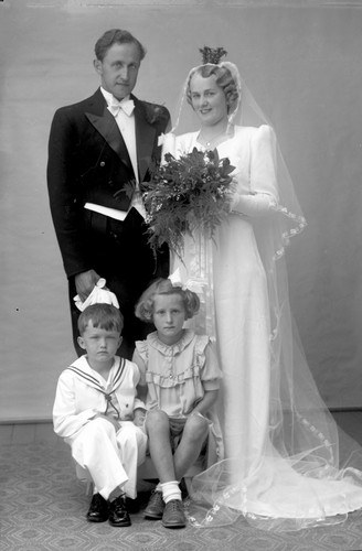 Schwarzes und weißes Foto einer Braut in einem weißen Kleid mit einem Strauß, Bräutigam, der neben ihr lächelt, und zwei Kindern, die vor ihnen auf dem Boden sitzen.