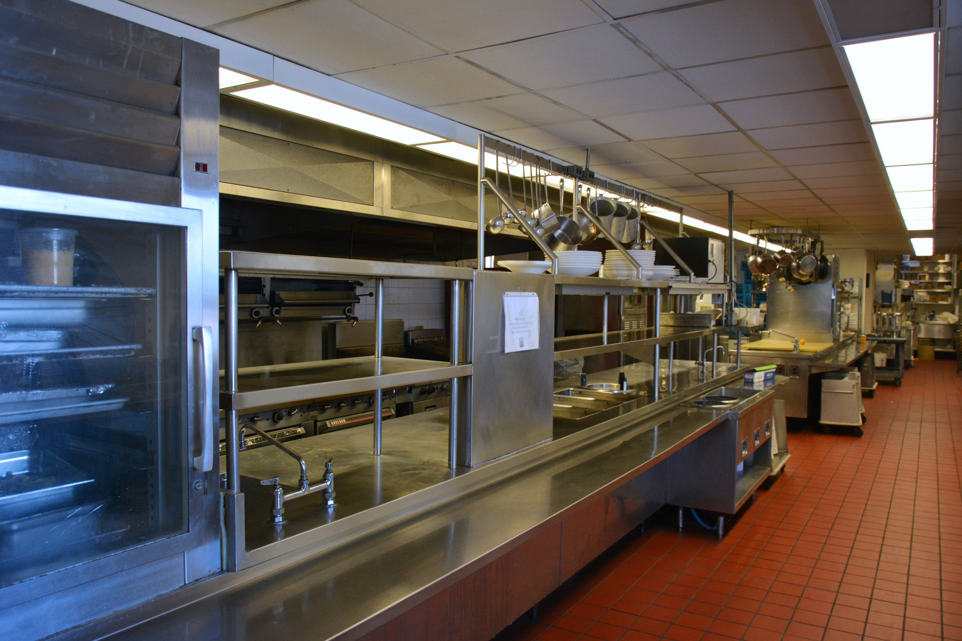Kommerzielle Küche mit Edelstahl-Theke, aufgehängten Utensilien, Spüle, Wasserhähnen, Schränken und diversen Gegenständen, die in Regalen unter elektrischem Licht angeordnet sind.