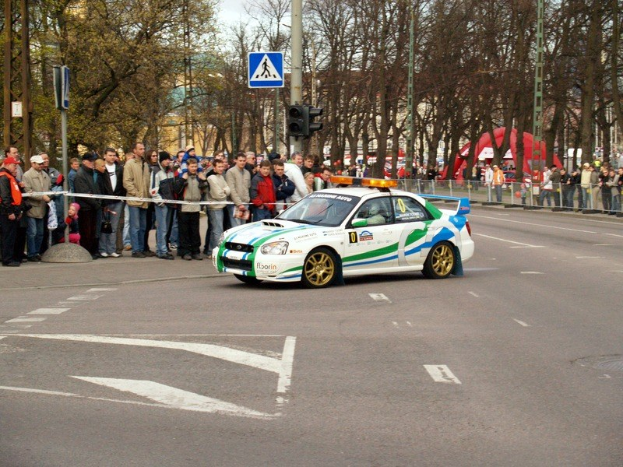 Ein Rallyefahrzeug fährt eine Straße mit Bäumen, Pfosten, Ampeln und Geländern entlang, während Zuschauer in Mützen von hinter Absperrungen zuschauen, mit Gebäuden und einem klaren blauen Himmel im Hintergrund.