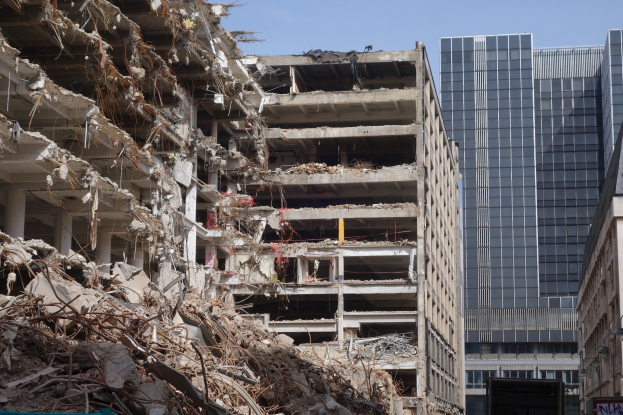 Ein großes Gebäude in der Innenstadt abgerissen, umgeben von intakten Gebäuden, Straßeninfrastruktur, Fahrzeugen, Bäumen und einem klaren Himmel.