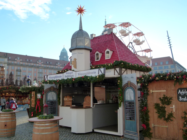 Ein kleines Gebäude mit einem roten Dach und einem Riesenrad davor, umgeben von Festdekorationen und Menschen, mit Gebäuden, Bäumen und einem bewölkten Himmel im Hintergrund auf einem Oktoberfest in München, Deutschland.