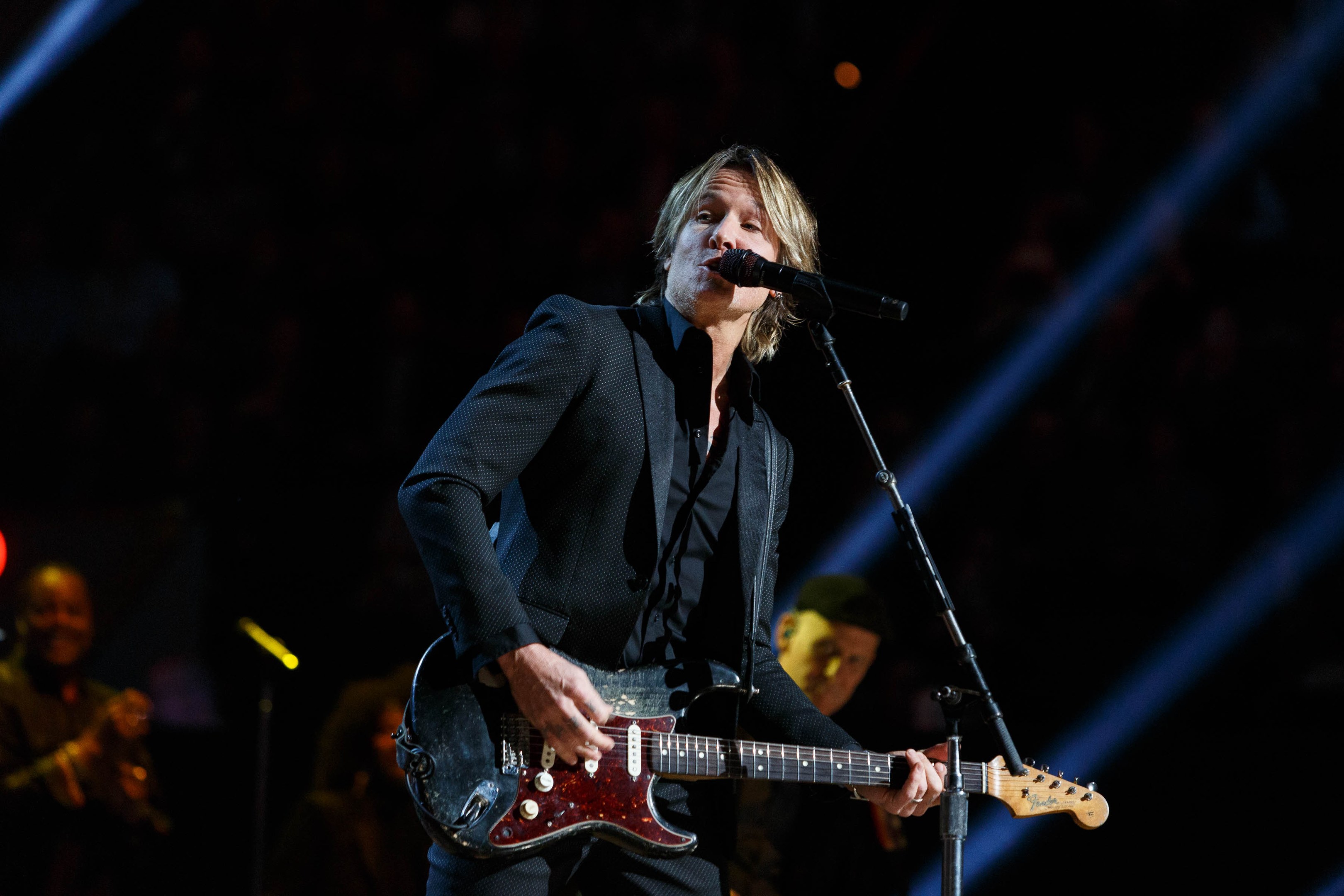 Keith Urban performs on stage at the Joint in Las Vegas, playing a guitar with a microphone in front of him and musicians in the background, under dark lighting.