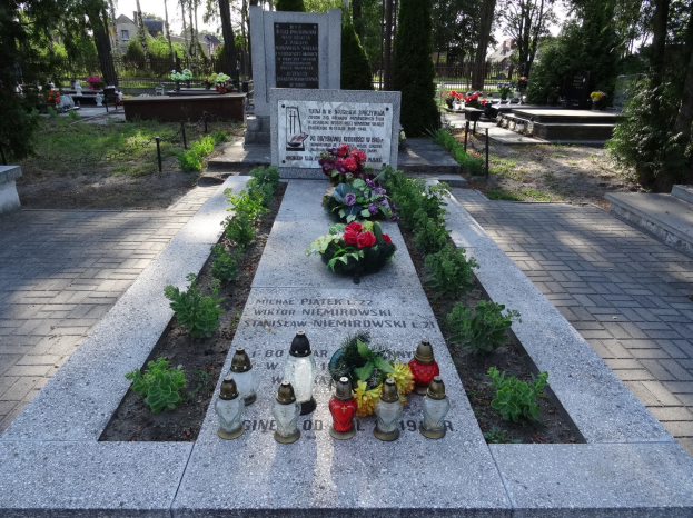 Denkmal in einem Park, gewidmet den Opfern der Tschernobyl-Katastrophe, umgeben von Blumen, Kerzen, Bäumen, Gebäuden und einem klaren blauen Himmel.
