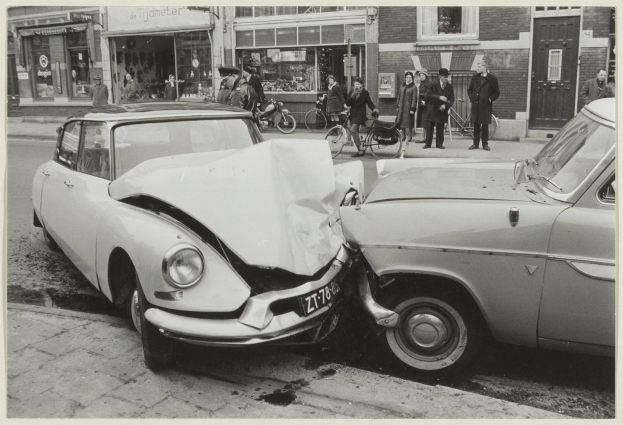 Schwarzes und weißes Foto von zwei beschädigten Autos am Straußerand mit Menschen und Fahrrädern in der Nähe, im Hintergrund sind Gebäude zu sehen.
