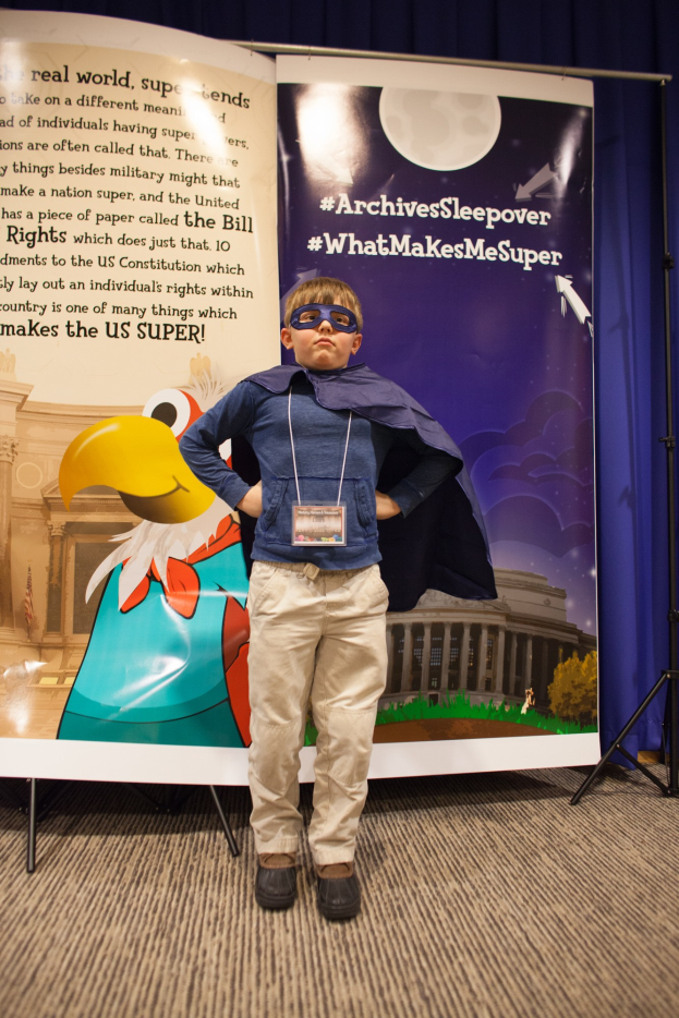 Ein junger Junge in einem Superheldenkostüm steht vor einer Banner mit Text und einem Vogelbild, mit einem Vorhang im Hintergrund und Schuhen.