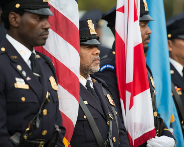 Eine Gruppe von Polizisten in Uniform und mit Mützen, die zusammenstehen und Fahnen halten, vor einem unscharfen Hintergrund.