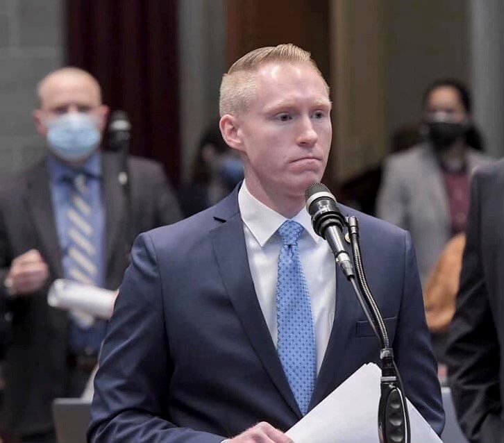 Ein Mann in Anzug und Krawatte spricht an einem Podium mit Papieren in der Hand, während hinter ihm ein paar Menschen stehen, nahe einer Vorhangwand.