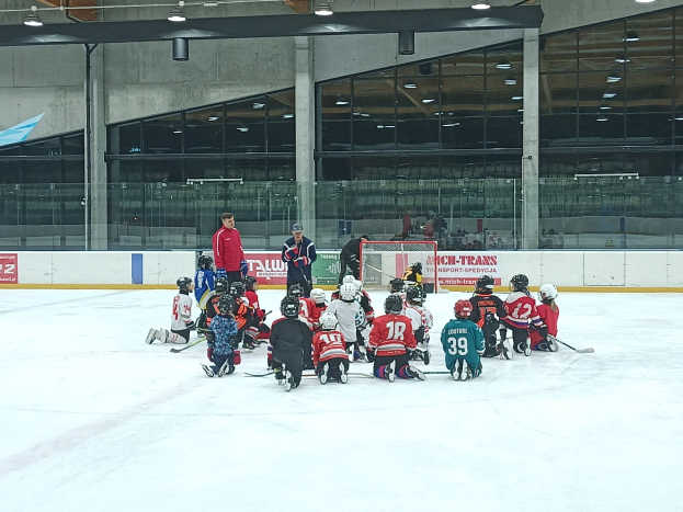 Kinder mit Helmen und Hockeystöcken auf einem Eisstadion sitzend, mit Glaswänden, Säulen, Deckenlampen und Informationsschildern im Hintergrund.