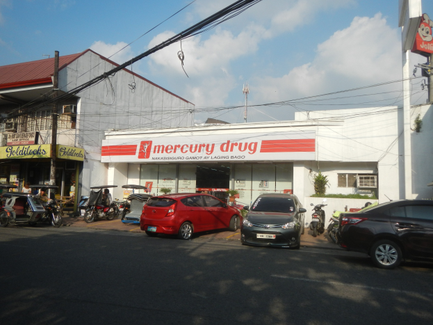 Eine belebte Straßenszene mit Fahrzeugen, Gebäuden mit Schildern, Strommasten mit Drähten, einer bewölkten Himmel und einer Mercury Drug Apotheke mit parkenden Autos davor.