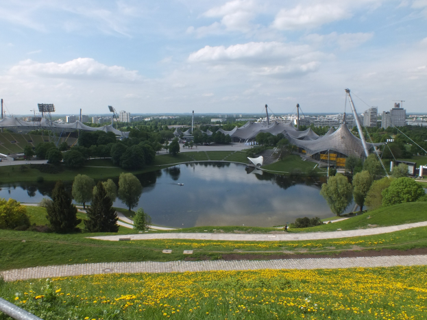 Ansicht des Olympiastadions von einem Hügel aus, mit einem Teich im Vordergrund, umgeben von Grünflächen und gelben Blumen, und Gebäuden im Hintergrund unter einem klaren blauen Himmel.