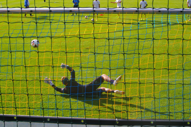 Ein Fussballspieler hält den Ball vor einem Tor, mit einem Netz im Vordergrund und einigen Menschen im Hintergrund, sowie mehreren Bällen auf dem Boden verteilt.