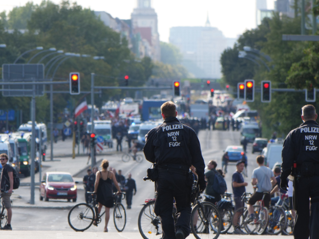 Zwei Polizeibeamte stehen an der Straße neben einer Gruppe von Menschen auf Fahrrädern, mit Fahrzeugen, Verkehrszeichen, Bäumen, Gebäuden und einem klaren blauen Himmel im Hintergrund.