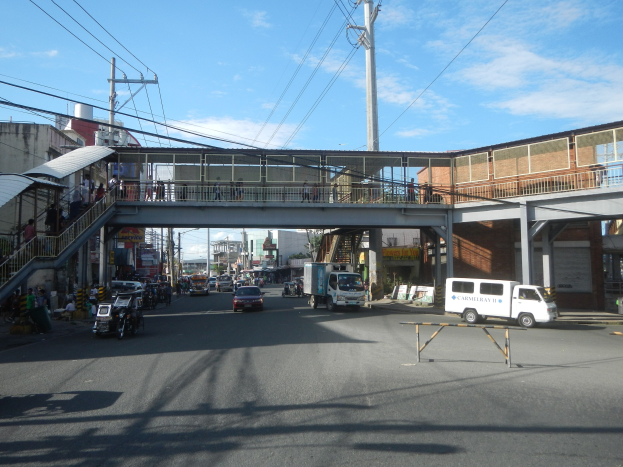 Eine Stadtstraße mit fahrenden Fahrzeugen, eine Fußgängerbrücke mit Menschen, Strommasten mit Drähten, Gebäude und ein bewölkter Himmel.