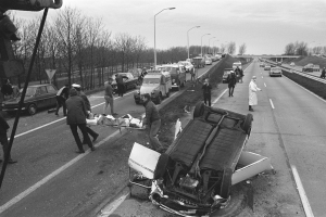 Schwarzes und weißes Bild eines umgestürzten Autos am Straßenrand mit beschädigten Fahrzeugen in der Nähe und Menschen, die sich darum versammeln, vor einem Hintergrund aus Laternenpfählen, Bäumen, einer Brücke und Himmel.