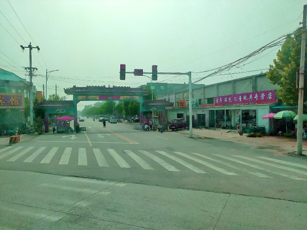 Stadtstraßenkreuzung mit Zebrastreifen, Ampel, Fahrzeugen, Strommasten, Gebäuden mit Beschilderung, Bäumen, Regenschirmen, ein Bogen im Hintergrund und sichtbarem Himmel.