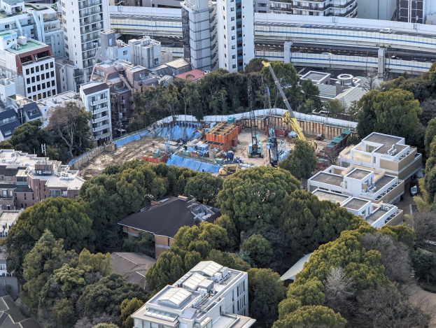Luftaufnahme einer Baustelle in einer Stadt mit Gebäuden, Bäumen, Fahrzeugen, Kränen und einer Brücke im Hintergrund, die verstreute Werkzeuge und Materialien zeigt.
