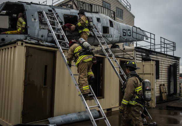 Feuerwehrleute in Uniform und Helmen arbeiten an einem Hubschrauber auf dem Dach eines Gebäudes, einer hält einen Sauerstoffzylinder, mit Leitern, einem Container, Bäumen und einem klaren Himmel im Hintergrund.