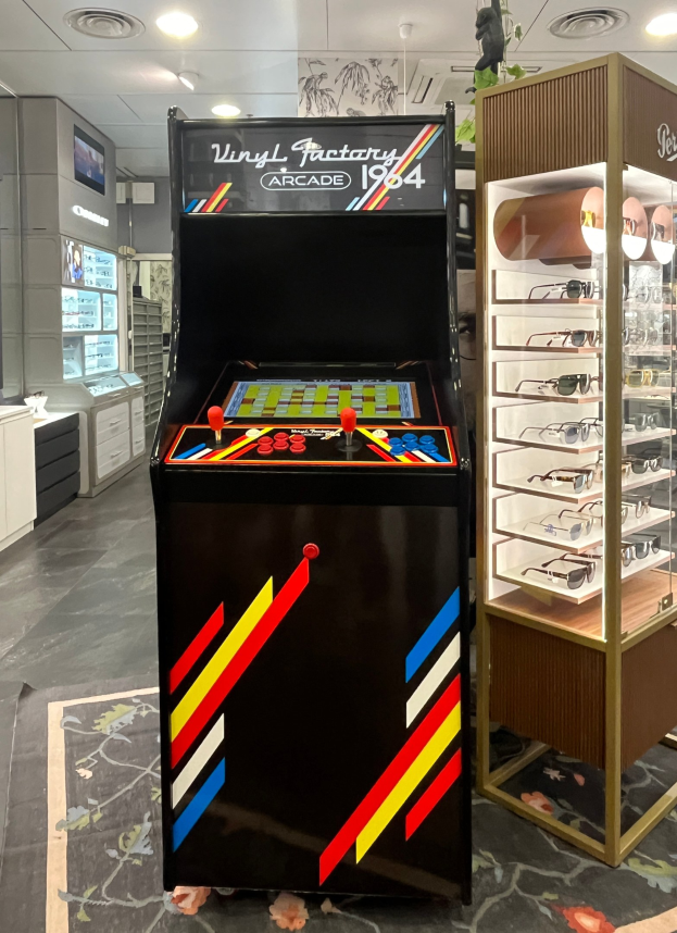 Ein Geschäft mit einem zentralen Retro-Arcade-Automaten, einer Brillenauslage auf der rechten Seite und einer retro-stilvollen Beleuchtung mit Schränken und einem Fernseher im Hintergrund.
