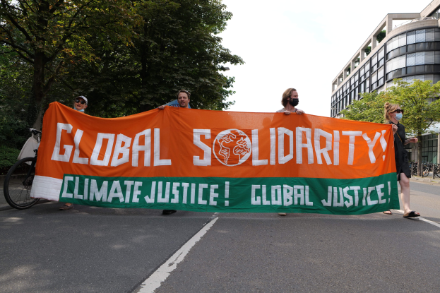 Gruppe von Menschen, die eine Straße mit einem 'Global Solidarity: Climate Justice'-Schild entlanggehen, während Fahrräder an der Straße geparkt sind und Gebäude im Hintergrund zu sehen sind.