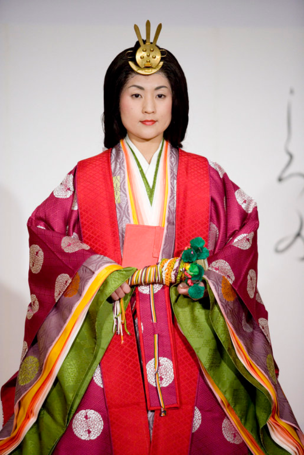 Eine Frau in einem traditionellen japanischen Kimono, das ein leuchtendes rotes und grünes Kleid mit aufwändigen Mustern zeigt, steht selbstbewusst auf einem Laufsteg, ihr Haar ist ordentlich zurückgebunden.