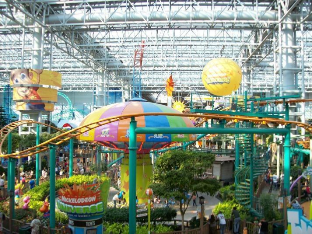 Ein großes Indoor-Wasserpark mit einer Achterbahn, umgeben von Menschen, Bäumen, Pflanzen und Deckenbeleuchtung mit Stangen.