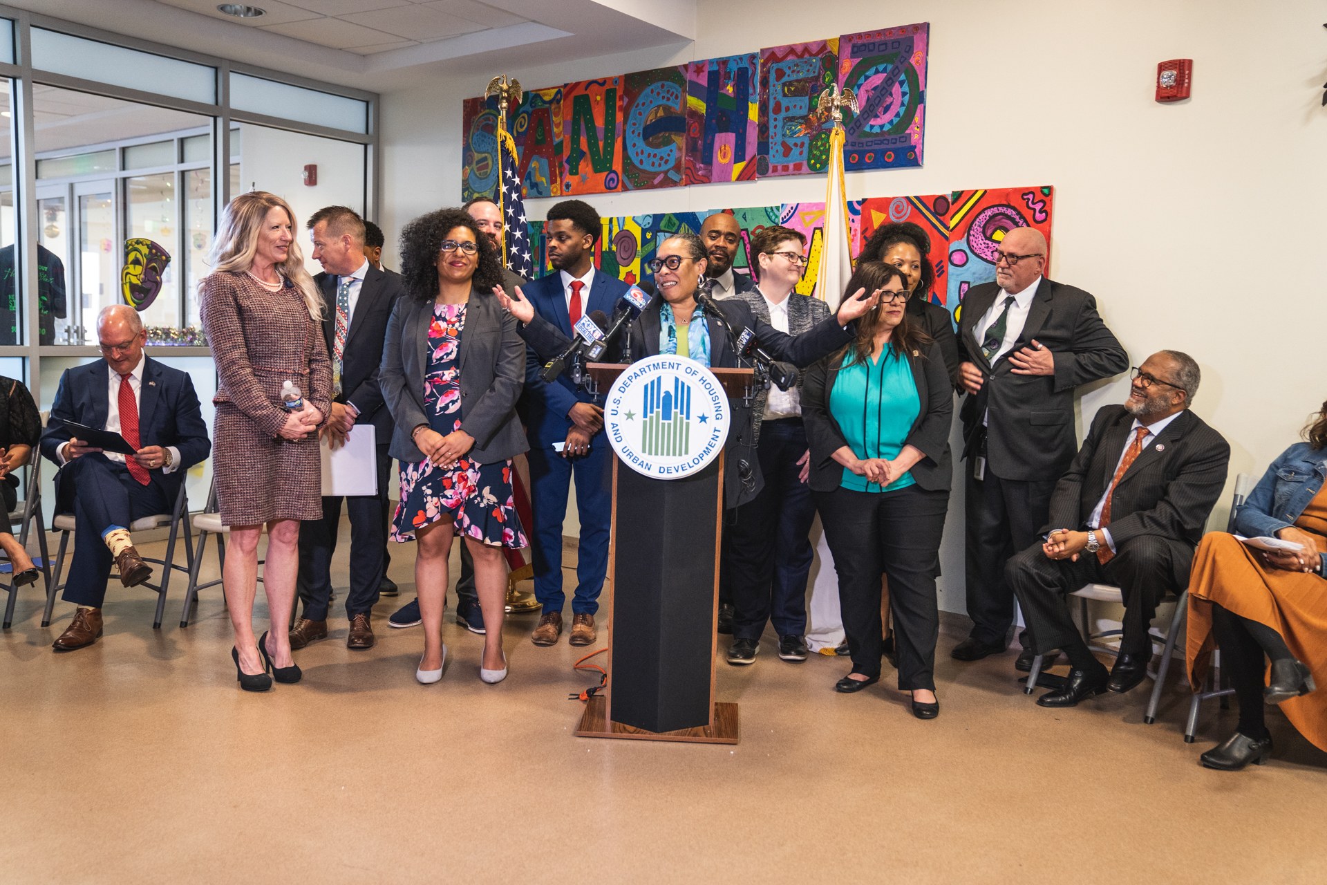 Gruppe von Menschen vor einem Rednerpult mit Mikrofon und einer Tafel mit der Aufschrift "Sancho Stadtrat tritt dem Stadtrat bei", mit sitzenden und stehenden Zuschauern, Flaggen, Feuerlöscher und Deckenleuchten im Hintergrund.