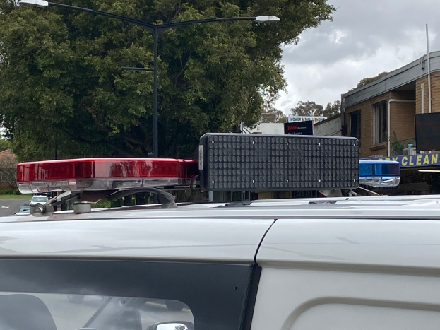 Ein Polizeiwagen mit eingeschaltetem Licht steht an der Straße neben Gebäuden, Bäumen, Pfählen und Schildern, unter einem bewölkten Himmel, wahrscheinlich nach einer tödlichen Schießerei an einer Schule in Brisbane.