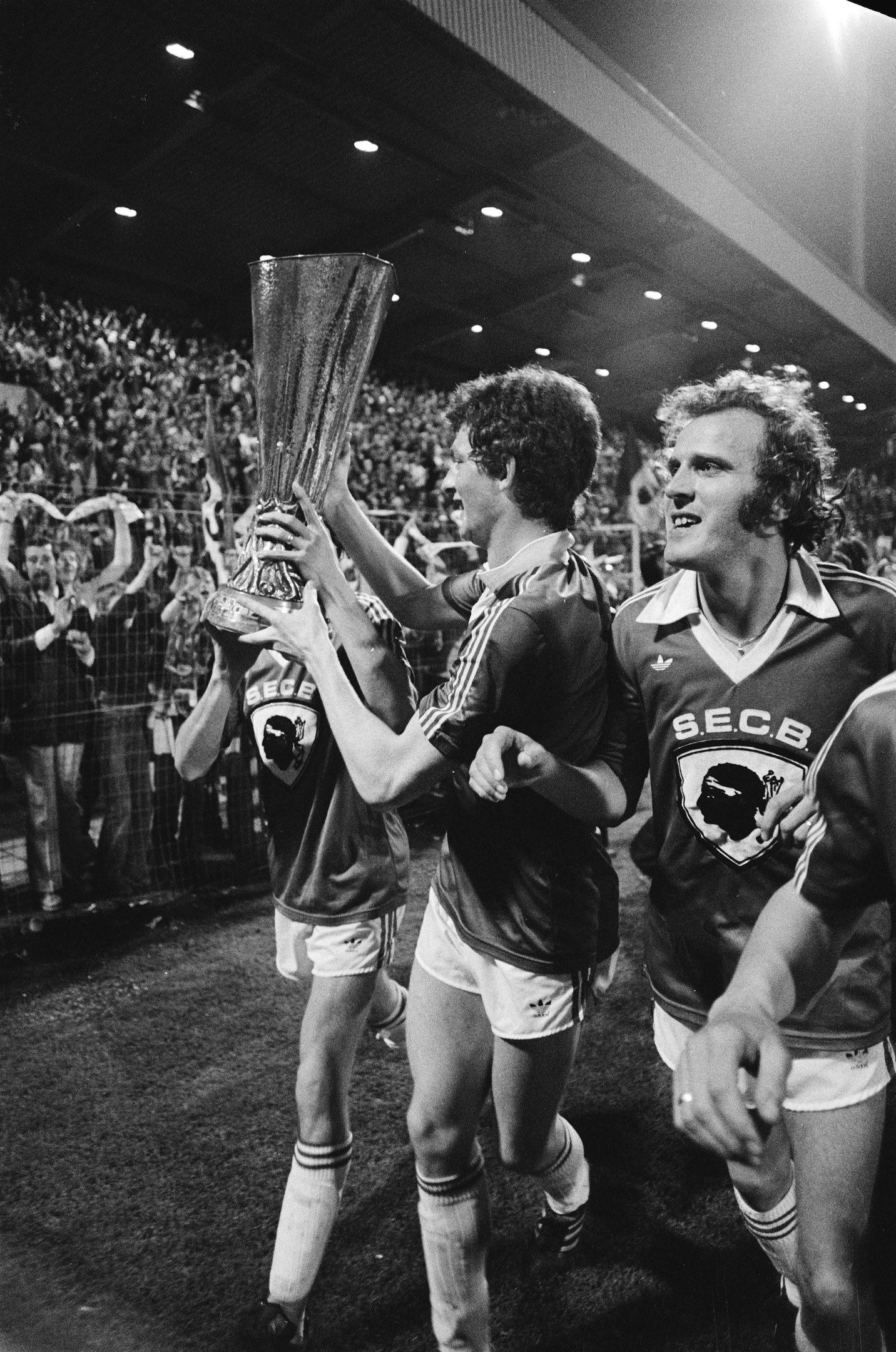 Schwarzes Bild von Fußballspielern, die mit einem Pokal auf einem Feld feiern, umgeben von Zuschauern und Stadionbeleuchtung.