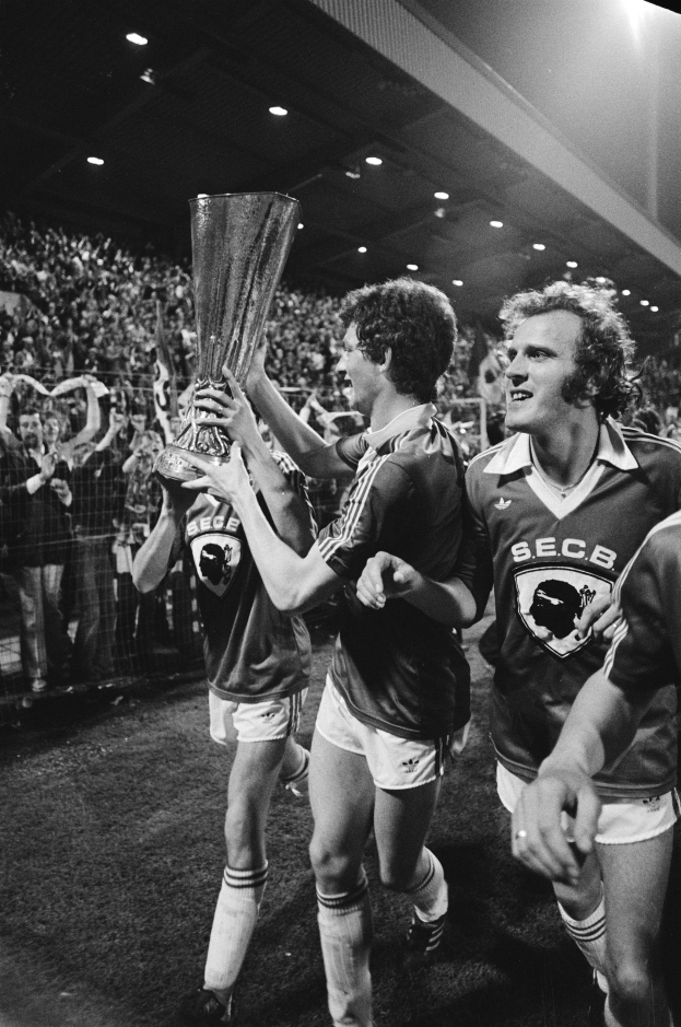 Schwarzes Bild von Fußballspielern, die mit einem Pokal auf einem Feld feiern, umgeben von Zuschauern und Stadionbeleuchtung.