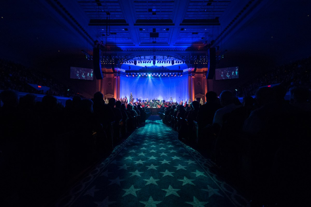 Großer Saal mit Sitzplätzen vor einer Bühne, auf der Musiker spielen, Decke mit Lampen und Fernsehern beleuchtet, dunkle Atmosphäre, die auf eine Konzerthalle hinweist.